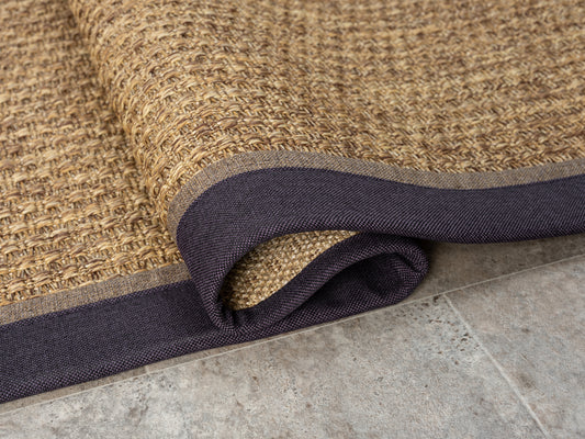 HALI VAGONU Mor Kahve Çift Renk Kumaş Bordürlü Sisal Halı'nın yuvarlanmış kenarını gösteren görüntü. El yapımı sisal halı, şık ev dekoru ve özel ölçü seçenekleri ile kişiselleştirilebilir. Bu dayanıklı ve evcil hayvan dostu halı, ofis için halı arayışında da göz önünde bulundurulmalı.