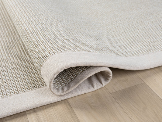 Krem ve açık tonlarda bir sisal halının, kenarındaki kumaş bordürle birlikte hafifçe yuvarlandığı bir açıdan görülüşü.