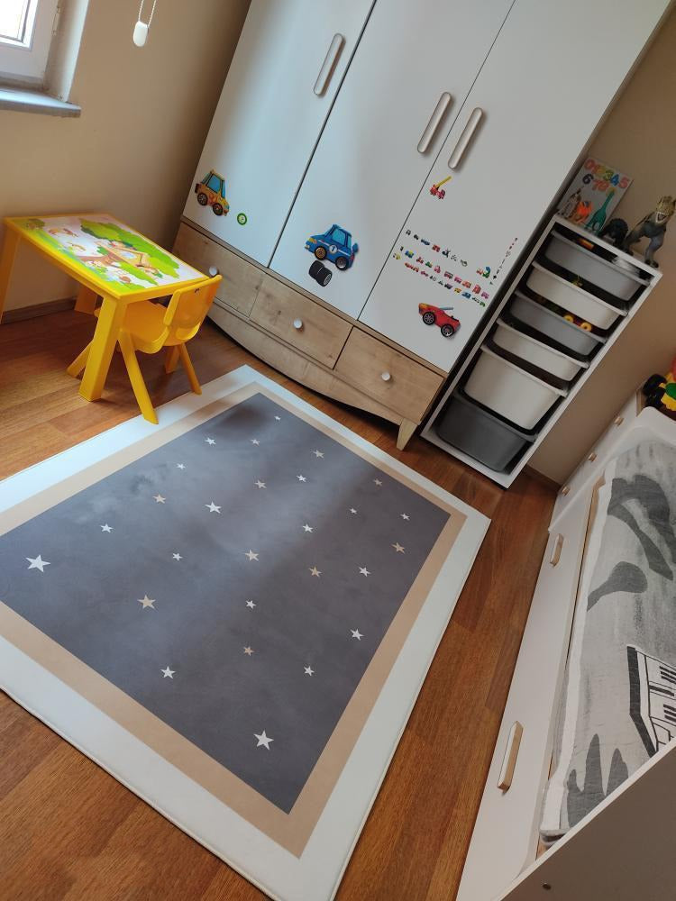 Gri çocuk halısı, etrafında beyaz çerçeve ve yıldız desenleri ile modern bir odada, sarı çocuk masası ve dolap ile birlikte yerleştirilmiş.