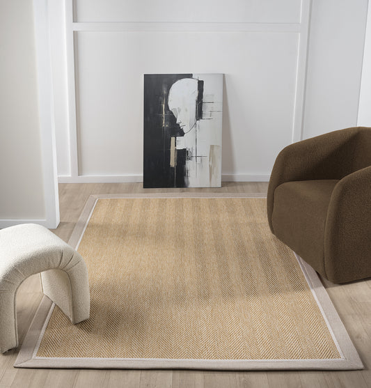 Modern bir oturma alanında yer alan balık sırtı desenli krem renkte sisal halı. Arka planda soyut bir tablo ve rahat bir koltuk görünmekte.