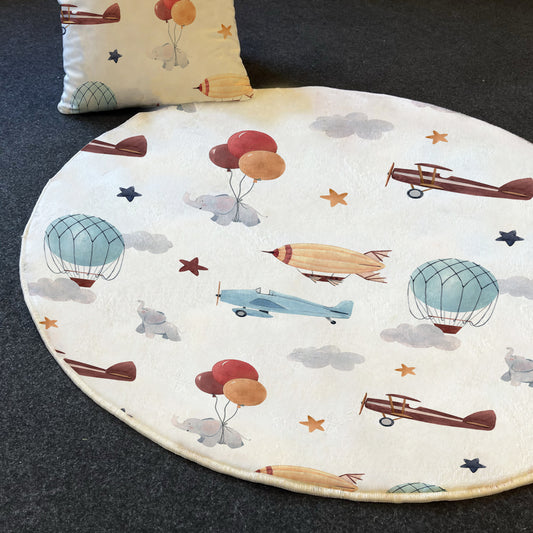 Yuvarlak şekilli uçaklar ve balonlar desenine sahip çocuk halısı, renkli figürlerle kaplı. Üstte bir yastık yer almakta.