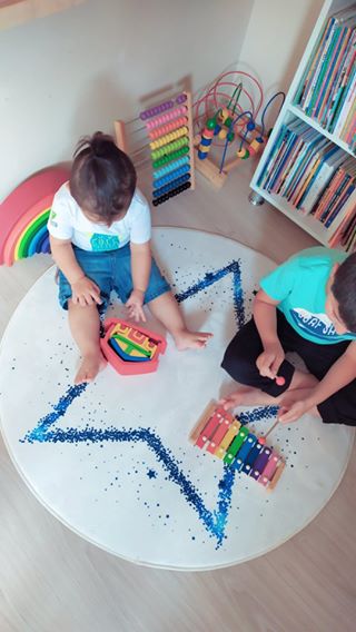 İki çocuğun, mavi yıldız desenli halının üzerinde oyuncak müzik aletleri ile oynadığı neşeli bir an.