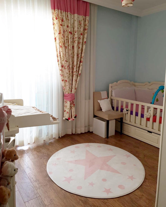 Bebek odasında yuvarlak pembe yıldız desenine sahip halı, ışık alan bir köşede serili. Odanın diğer köşesinde bebeğin beşiği ve şirin perdeler yer alıyor.