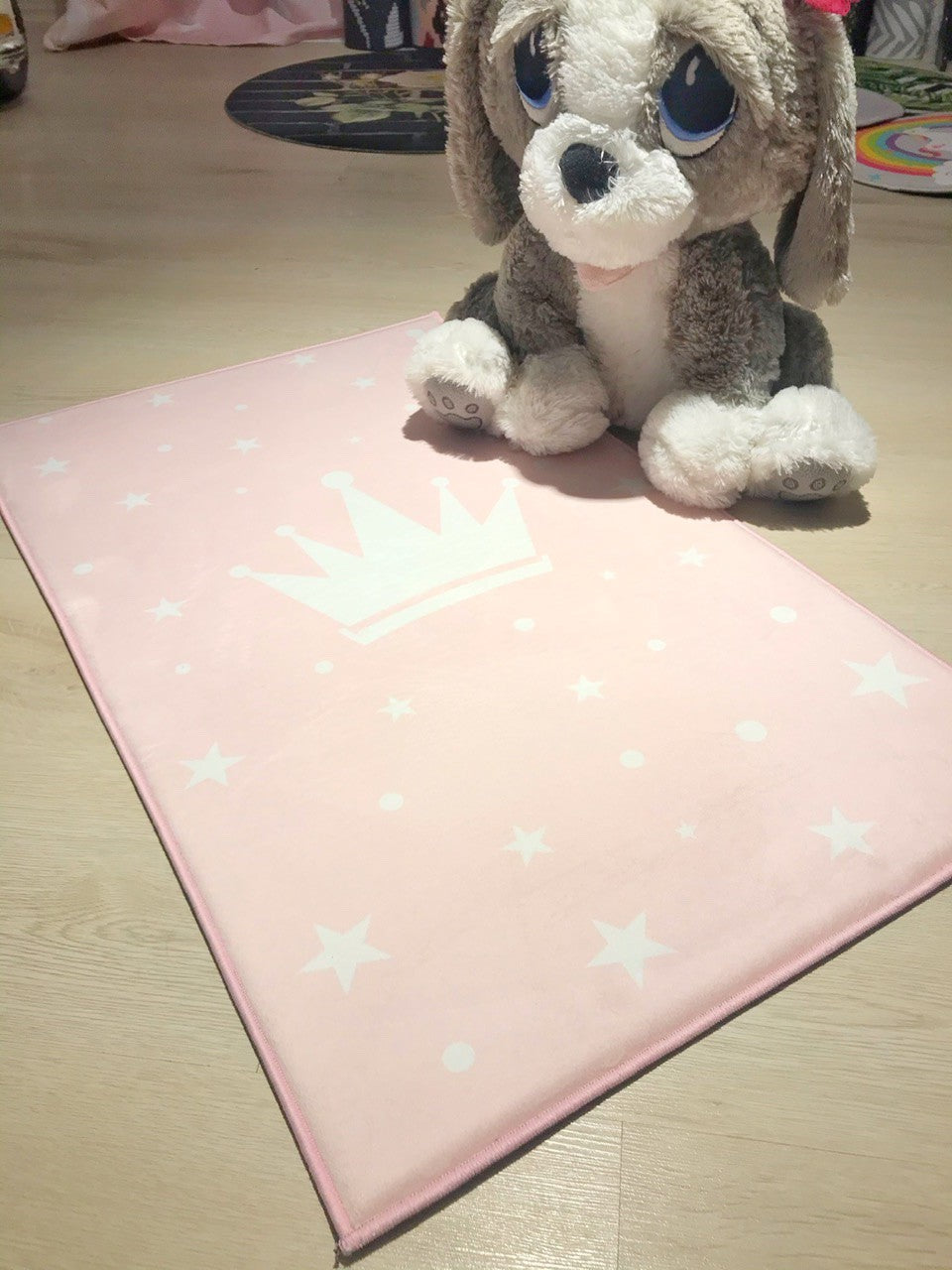 Pembe renkli, taç desenli ve yıldızlı çocuk odası halısı, halının üzerinde oturan sevimli peluş köpek oyuncak ile birlikte gösterilmektedir. Halı, yumuşak dokusu ile oyun alanları için ideal bir seçimdir.