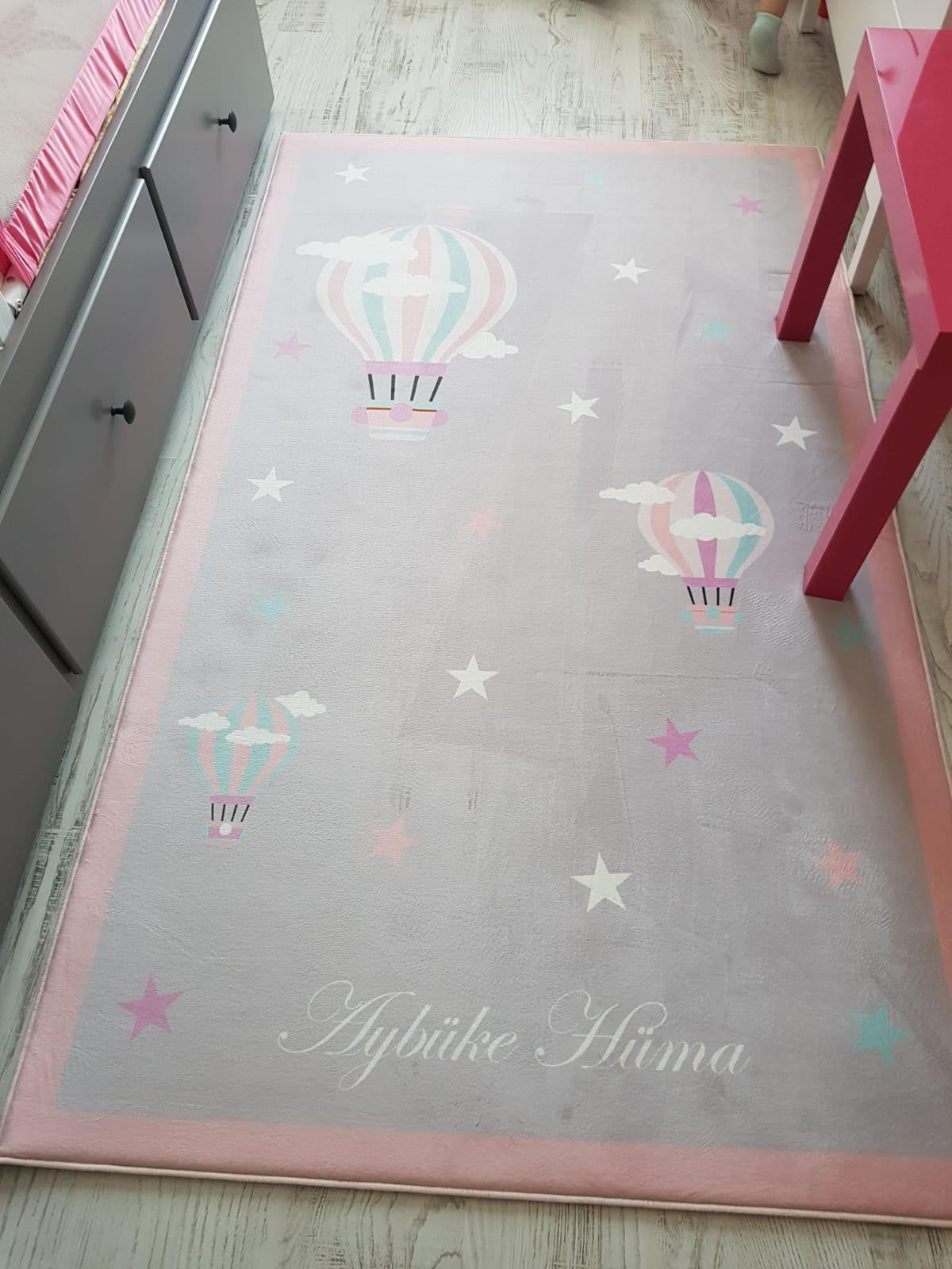 Yıldız desenli ve uçan balon motifli çocuk halısı, pembe kenarlarla çevrili, çocuk odasında yumuşak ve rahat bir alan sunuyor.