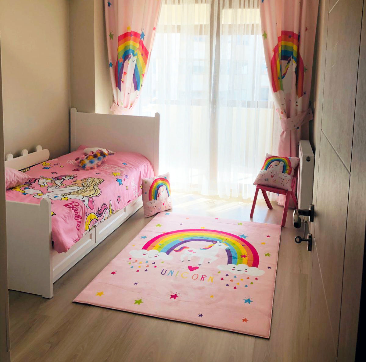 Işık dolu bir çocuk odasında, pembe unicorn desenli halı, beyaz yatak ve diğer dekoratif öğelerle birlikte yer alıyor.