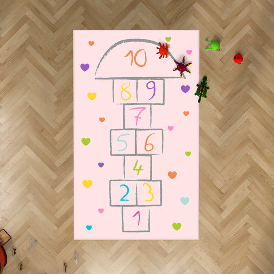 Pembe çocuk halısının üzerinde renkli kalpler ve rakamlar ile tasarlanmış sek sek oyunu. Halı yumuşak dokusu ile çocukların oyun alanında rahat bir kullanım sunar.