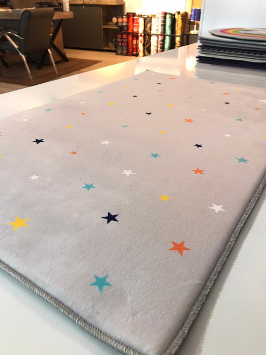 Düz açık gri yüzeyde çeşit çeşit renkli yıldızlarla süslenmiş halı, modern bir ortamda sergileniyor.