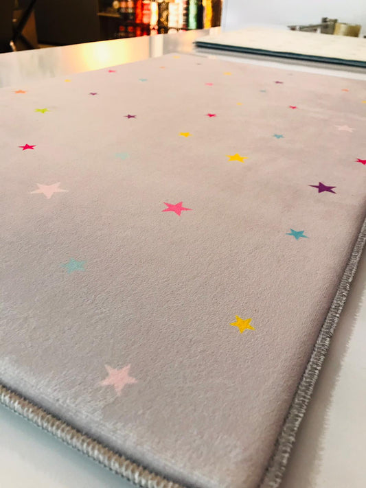 Açık gri bir halı, üzerinde farklı renklerde yıldızlar yerleşmiş. Halı, kenardan biraz yükseltilerek çekilmiş, dokusu ve desenleri belirgin şekilde gözüküyor.