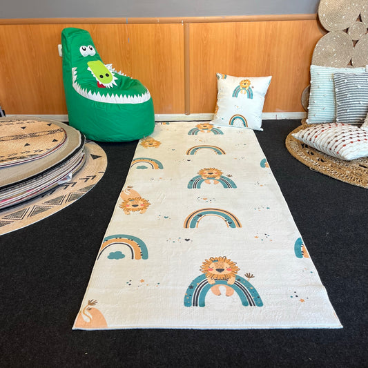 Çocuk odasında yeşil bir pufun önünde yer alan aslan ve gökkuşağı desenli halı. Halının yanında uyku yastığı ve yuvarlak halı yer almakta.
