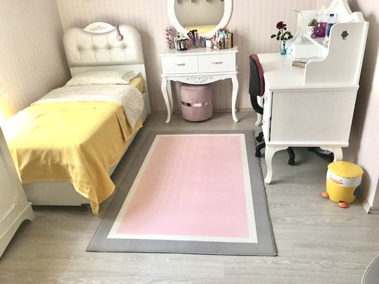 Yatak odasında, sarı yastıklı yatak ve beyaz mobilyalarla birlikte yumuşak gri pembe çocuk halısı serili.