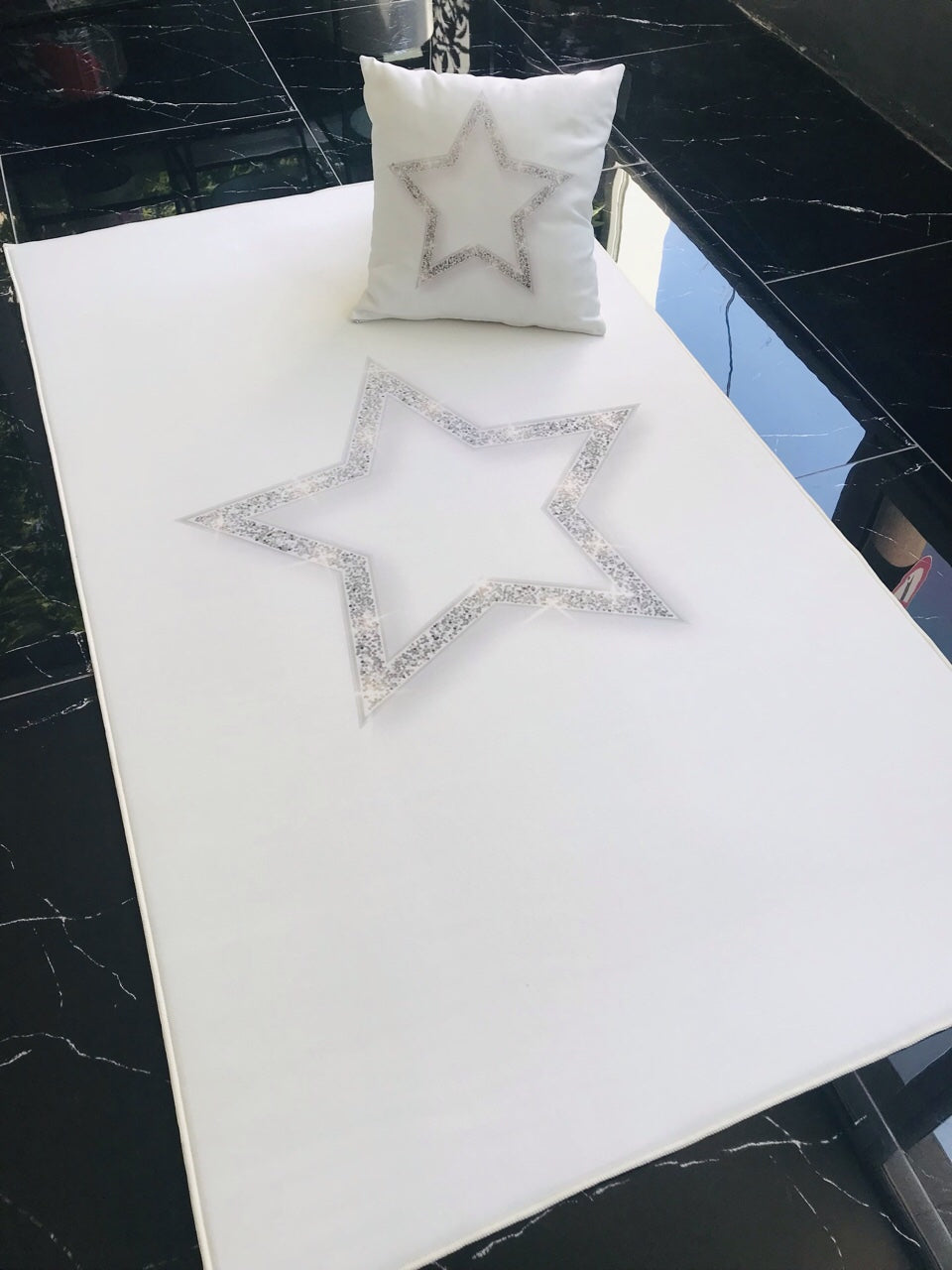 Gümüş yıldız desenli halı ve üzerine konulmuş yastık, beyaz zemin üzerinde modern bir görünüme sahip. Halının çevresinde şık bir dikiş detayı bulunuyor.