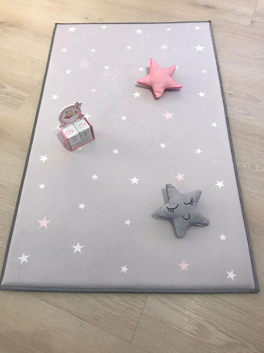 Çocuk odasında pembe ve gri yıldızlarla süslenmiş açık gri halı, üzerinde sevimli yıldız oyuncaklar ve bir takvim yer alıyor.