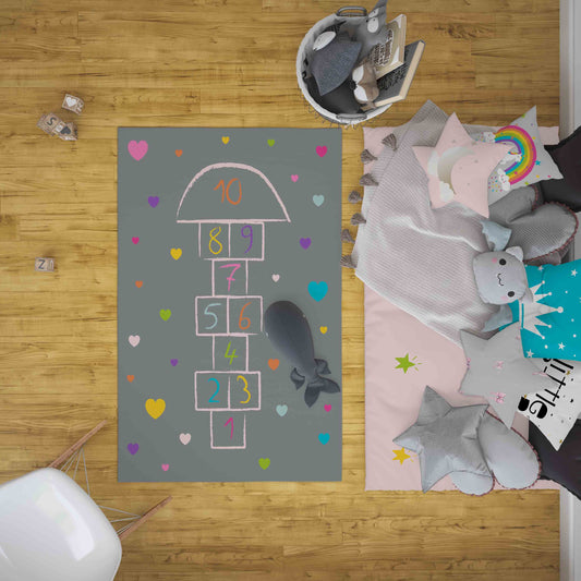 Halı Vagonu HV342 Seksekli Kalpli Gri Çocuk Odası Halısı'nın kullanıldığı bir odanın üstten görünümü. Halının üzerinde rengarenk kalp ve rakamlar var, etrafta oyuncaklar ve yastıklar serili.