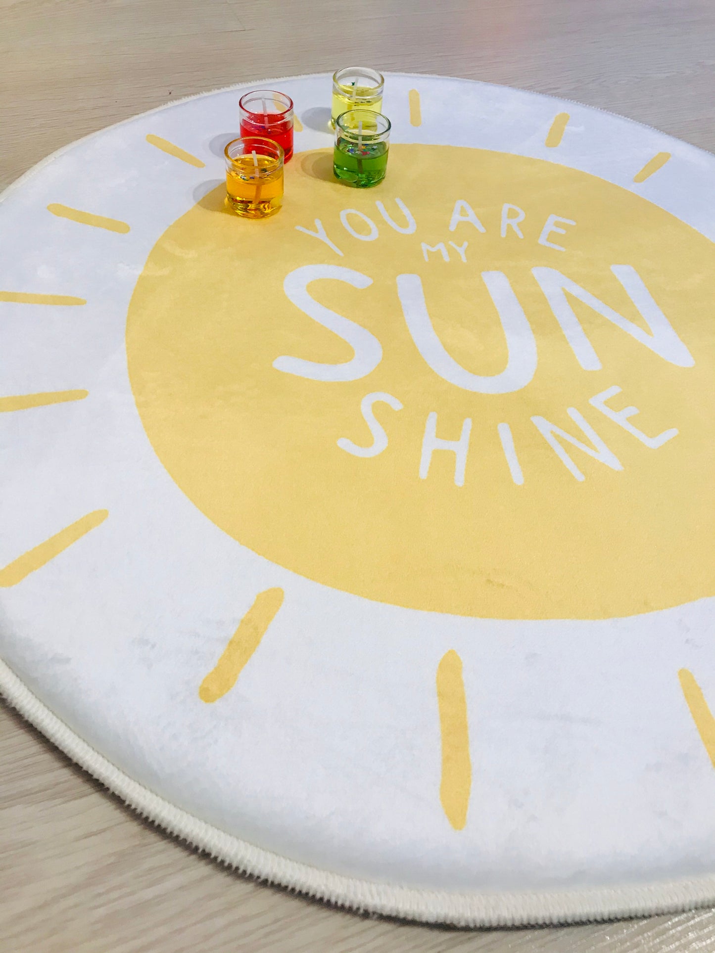 Sarı ve beyaz renklerde, 'You Are My Sunshine' yazılı yuvarlak halı, üzerinde renkli bardaklarla birlikte sunulmuş, eğlenceli bir oyun alanı imajı yaratıyor.