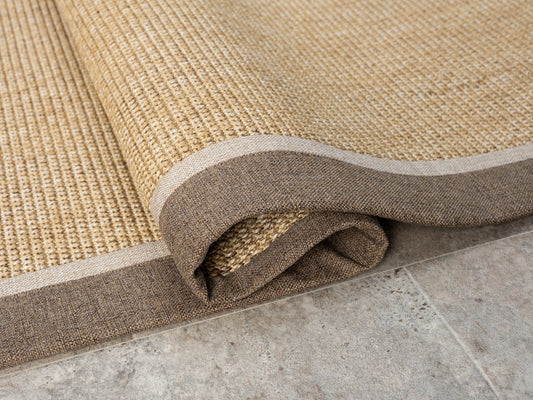 Yuvarlanmış el yapımı sisal halının üst kısmı, kumaş bordür detaylarıyla birleşmiş. Halının dokusu ve kenarındaki çift renkli bordür net bir şekilde seçiliyor.