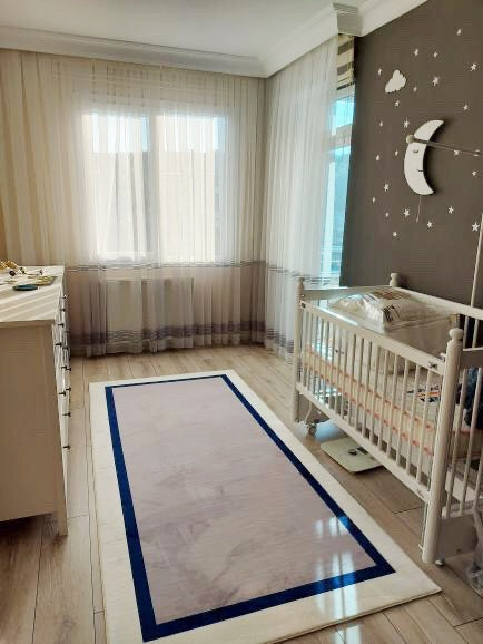 Aydınlık bir bebek odasında, açık renkli bir çocuk halısı. Halı, gri lacivert beyaz çerçeve ile tasarlanmış. Odanın dekorasyonunu tamamlayan doğal ışık alan pencereler ve beyaz bebek yatağı dikkat çekiyor.