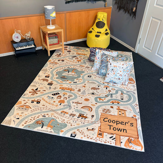 Cooper's Town temalı çocuk halısı, şirin desenleriyle birlikte bir odada yer alıyor. Beyaz zemin üzerine pastel renklerle çizilmiş yollar, evler ve hayvan figürleri görünmekte. Halının yanı başında sarı bir bean bag ve küçük bir masa var.