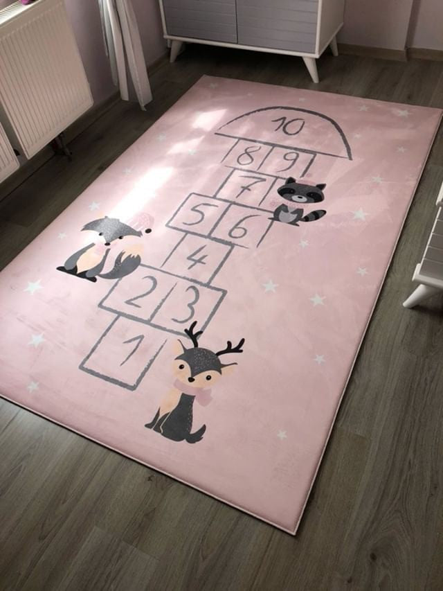 Yumuşak pembe zemin kaplaması üzerinde, çocuklar için tasarlanmış oyun alanını temsil eden hopscotch oyun şemasını gösteren bir halı. Halının köşelerinde sevimli kuş ve geyik figürleri yer alıyor, çocukların oyun alanını renklendiren yıldız deseni ile tamamlanıyor.